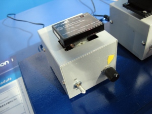 オムロンの20×20×4mm/重さ3.7gの超小型の発電体を2個組み合わせた発電モジュール「BP-02」。テスト装置に上に載った黒い箱がそれ。38×54×10mm/22gの寸法から5.2V/0.1mW(暫定値)の電力が得られるという