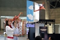 「ORIZURU」を手に飛行させるコンパニオンの向こう側で真剣な表情の男性が、開発に協力した超小型飛行体研究所の宗像俊龍氏