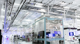 A modern industrial facility interior with advanced machinery and bright lighting. The equipment includes various computer monitors and high-tech apparatus, surrounded by clean, organized workspaces. People in protective suits work in the background. (Intel.com)